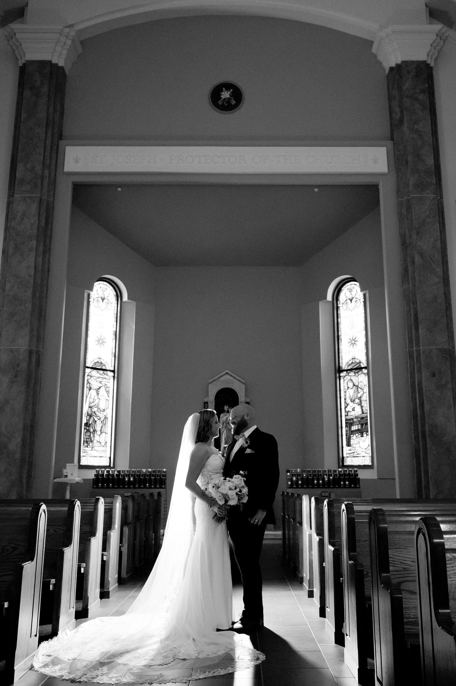 Black and white photo of bride and groom kissing at church altar, flanked by pews and arched stained glass windows.