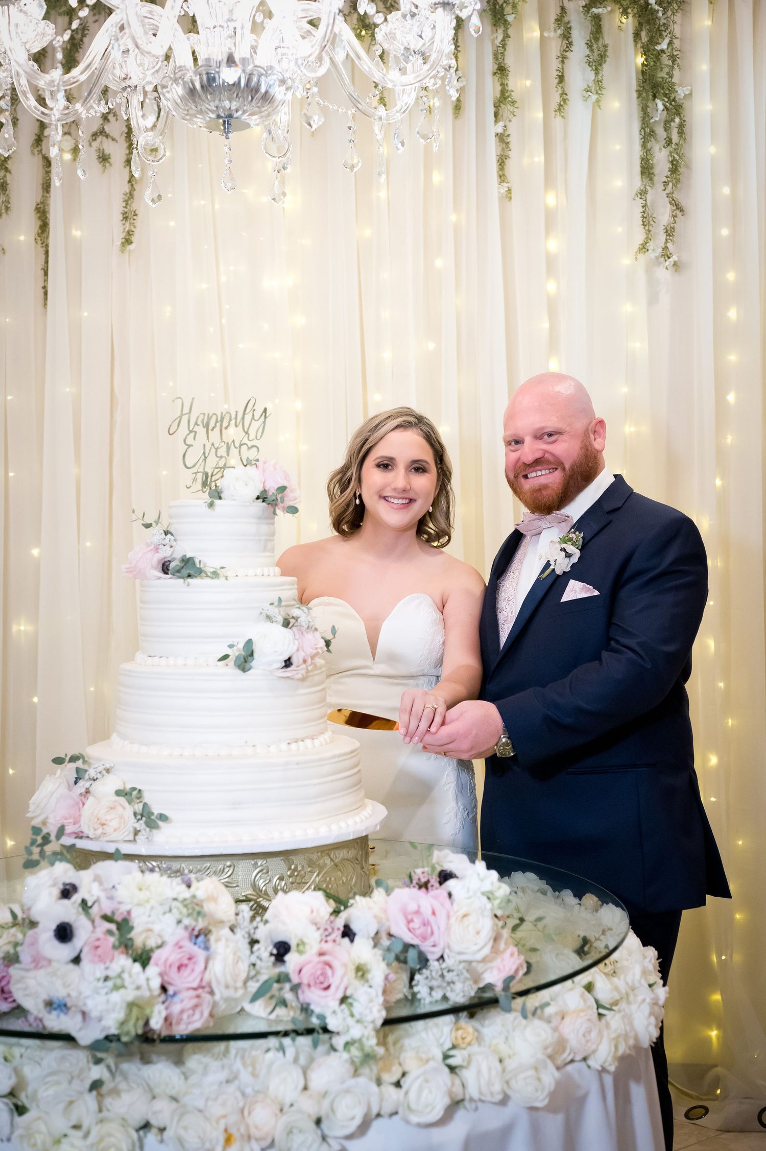 Bride and groom cutting a white four-tier wedding cake adorned with flowers and a Happily Ever After topper.