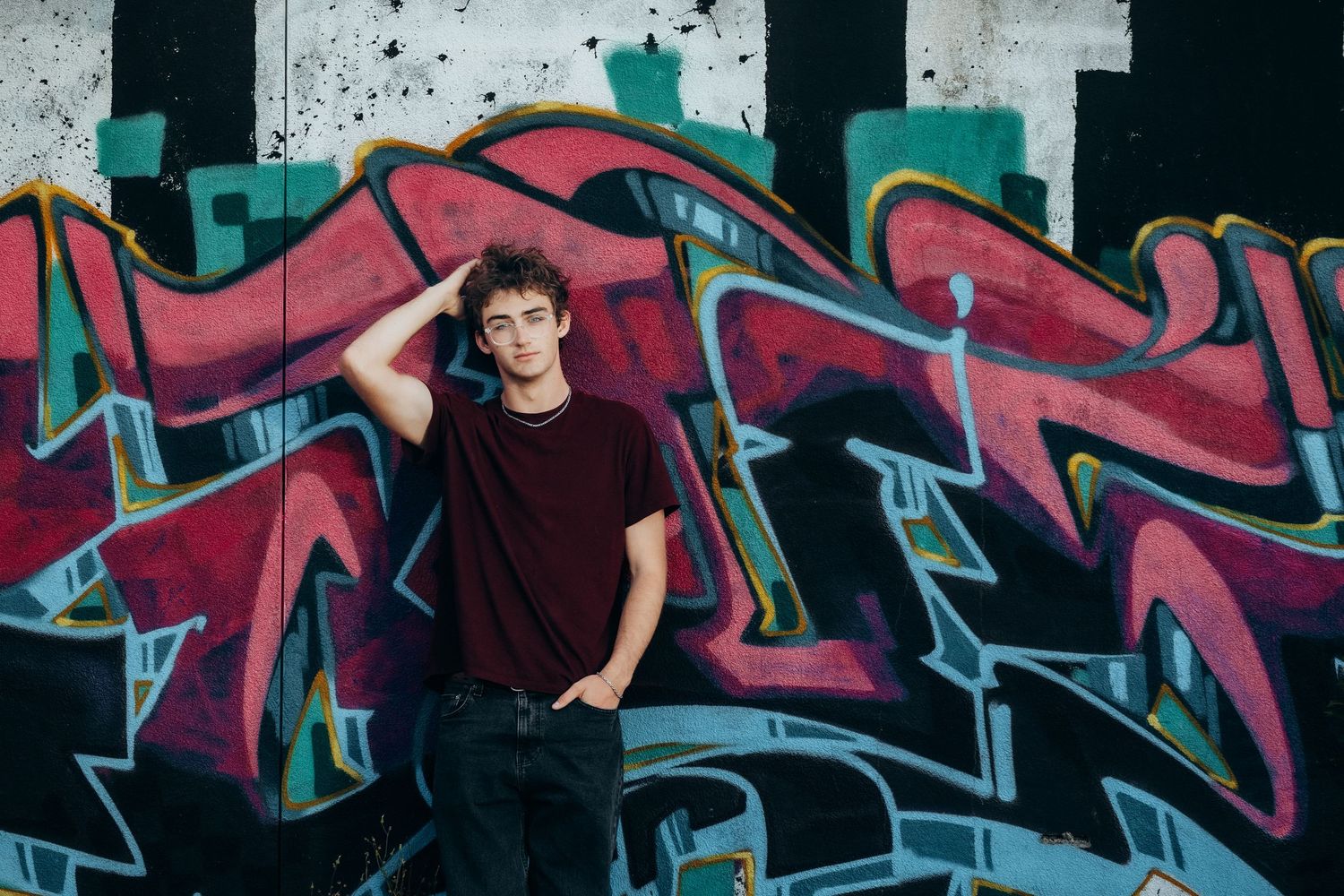 Young man in dark red t-shirt poses confidently against vibrant colorful graffiti wall outdoors.