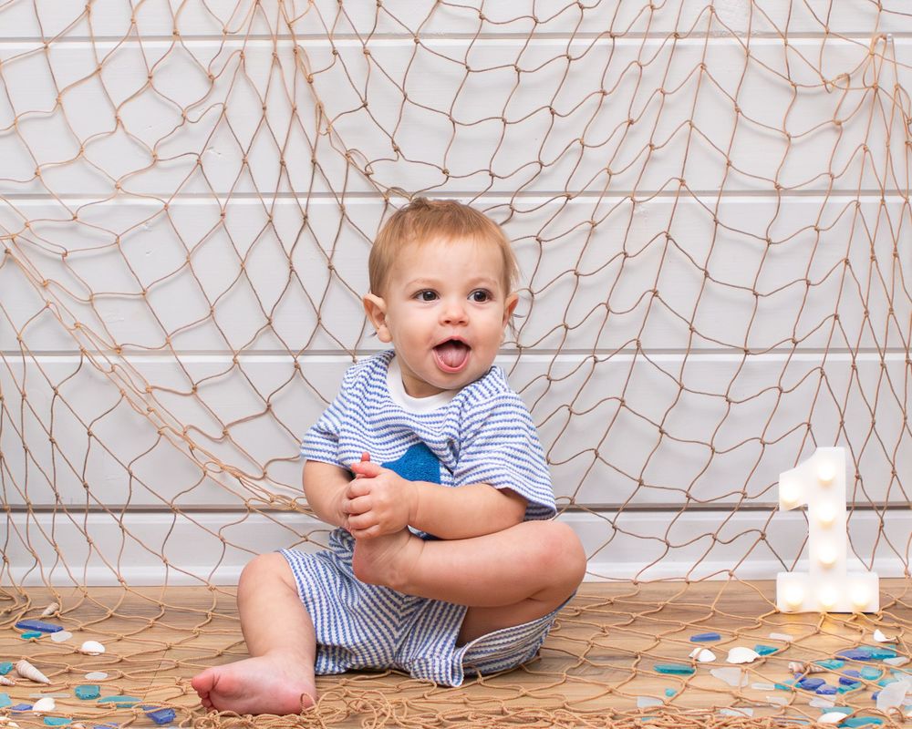 Smiling baby boy in striped outfit sitting on sandy floor by fishing net with number 1, celebrating first birthday.