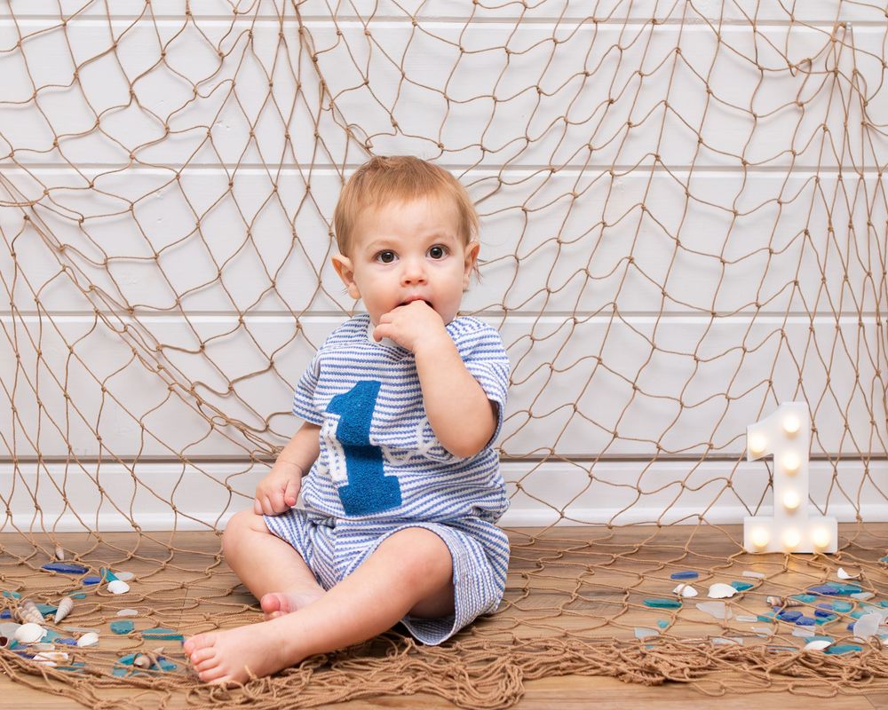 Baby boy in striped outfit sits by fishing net backdrop celebrating his first birthday.