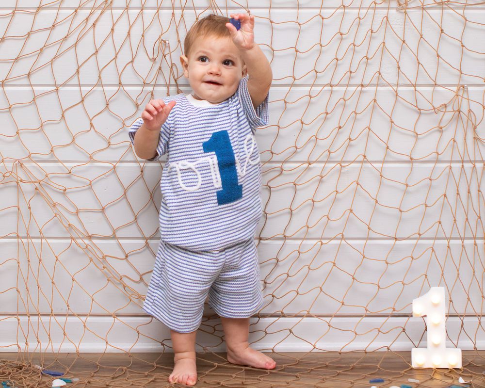 Baby boy in striped blue outfit with number 1, standing by fishing net backdrop celebrating first birthday.