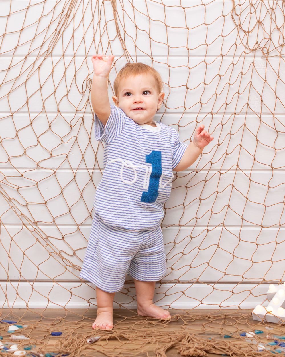 Baby boy in striped outfit with number 1 stands holding a fishing net backdrop at his first birthday.