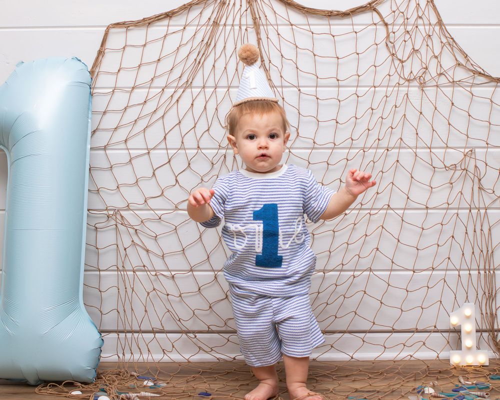 Baby boy in striped outfit with number 1 shirt celebrates first birthday beside blue balloon and fishing net backdrop.