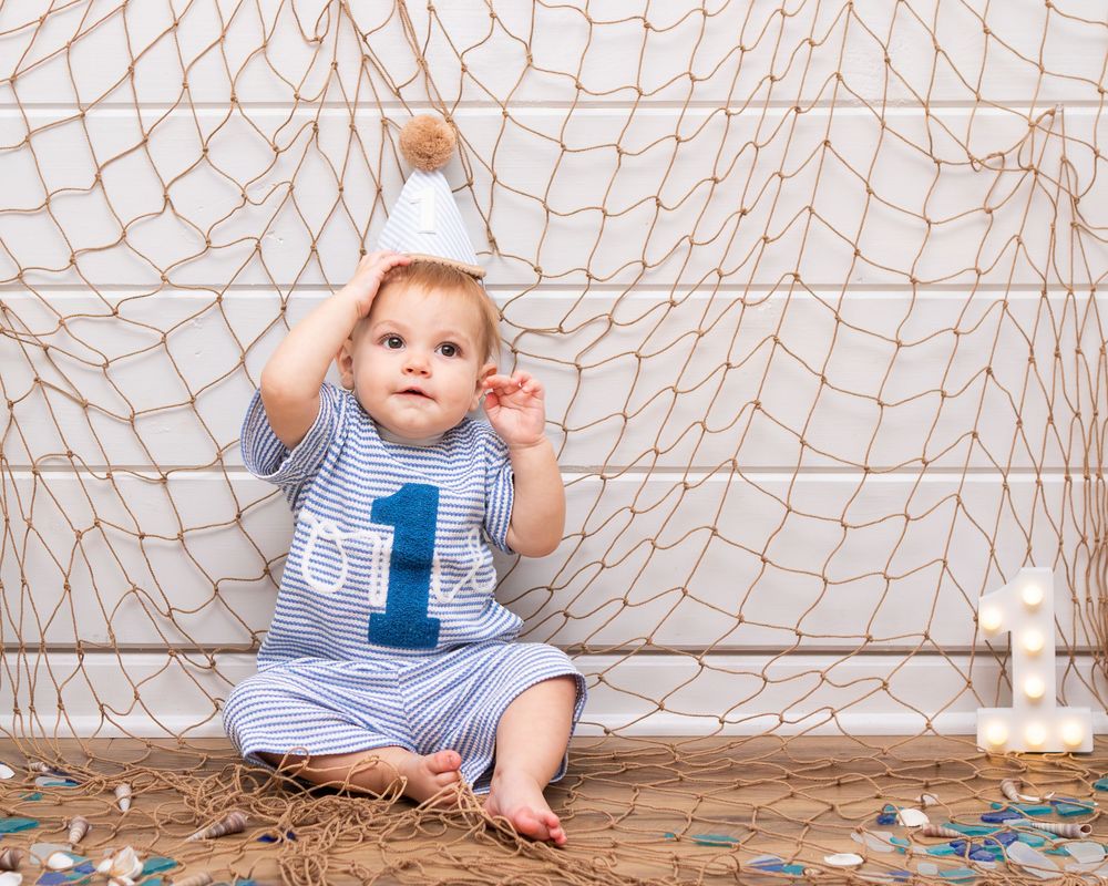 Baby boy in striped romper with number 1, wearing party hat, sitting by fishing net backdrop for first birthday photos.