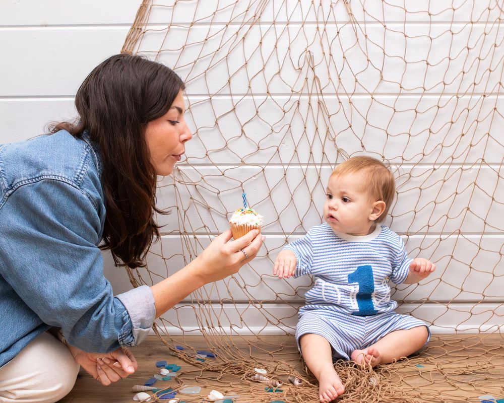 Mother blowing out candle on cupcake for baby's first birthday celebration with fishing net backdrop.