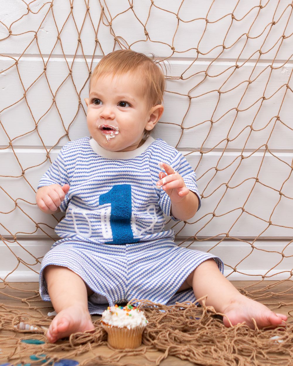 Baby in striped romper with number 1 eating birthday cupcake, sitting on fishing net for first birthday cake smash.