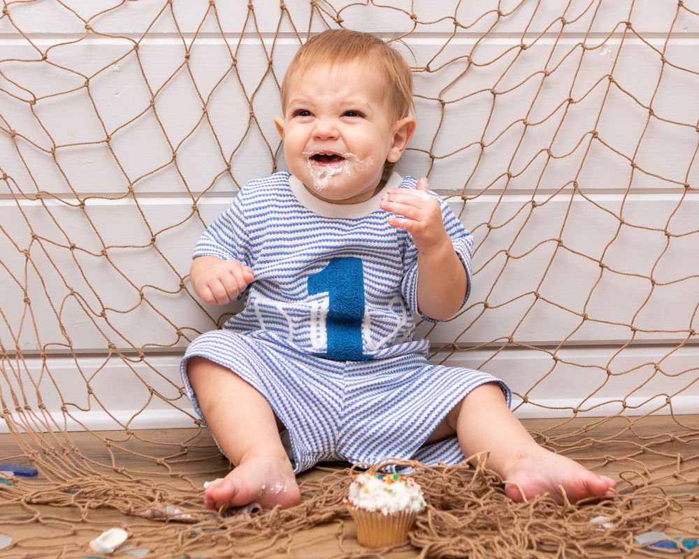 Happy baby boy celebrating first birthday, laughing with frosting on face next to cupcake and fishing net backdrop.