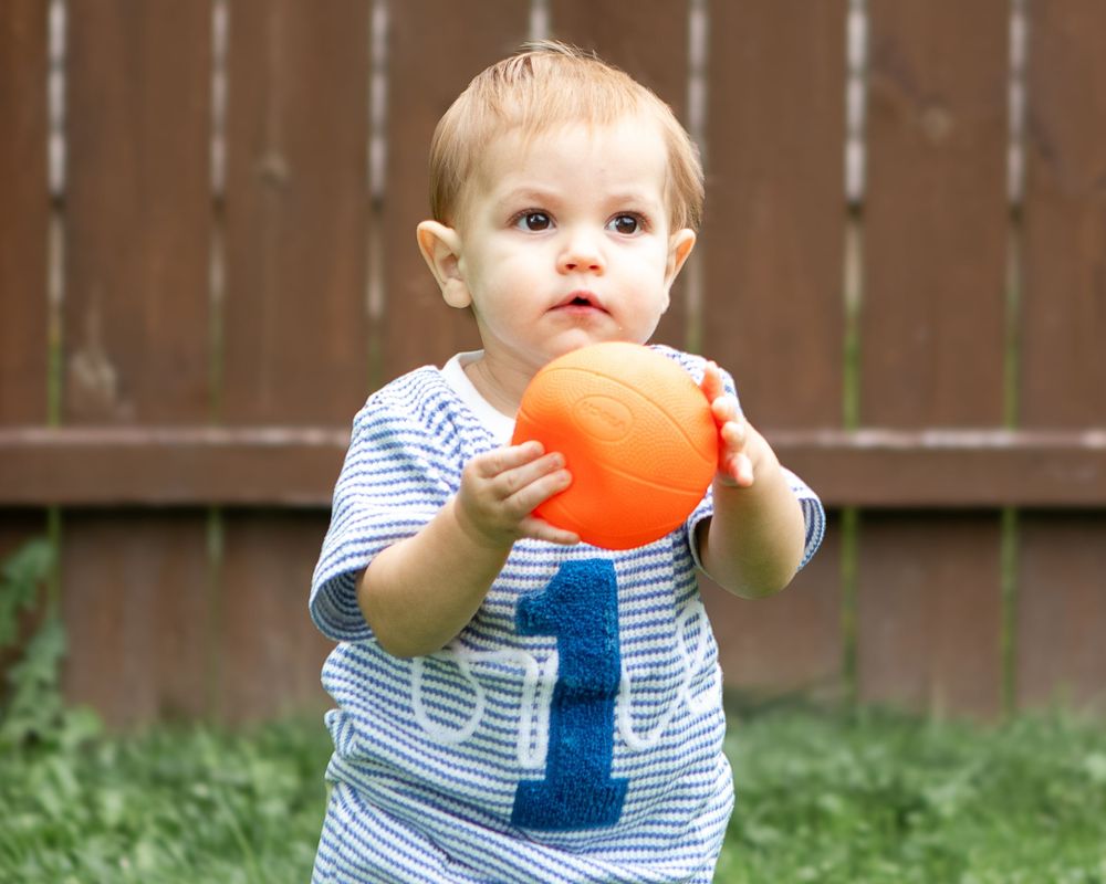Toddler boy in striped shirt holding an orange ball outdoors in a backyard garden setting.