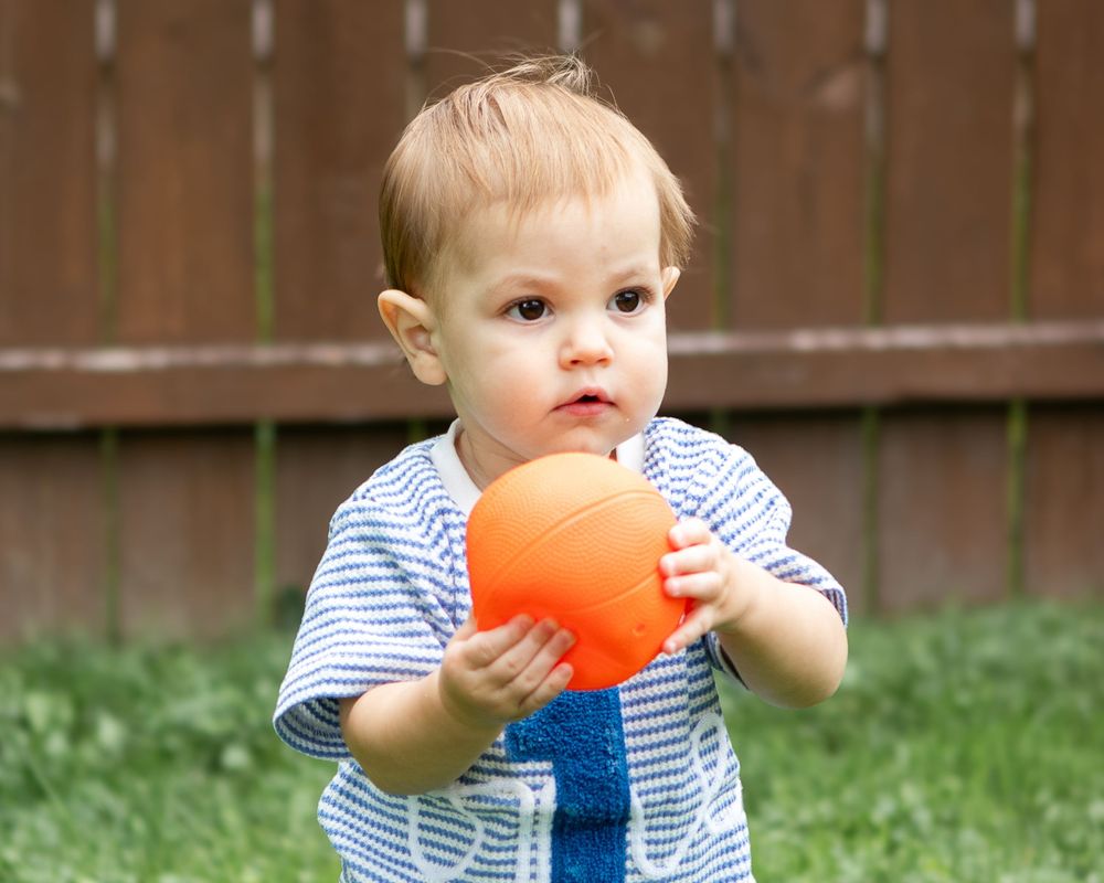 Toddler boy in blue striped shirt holds an orange ball while standing on green grass outdoors.
