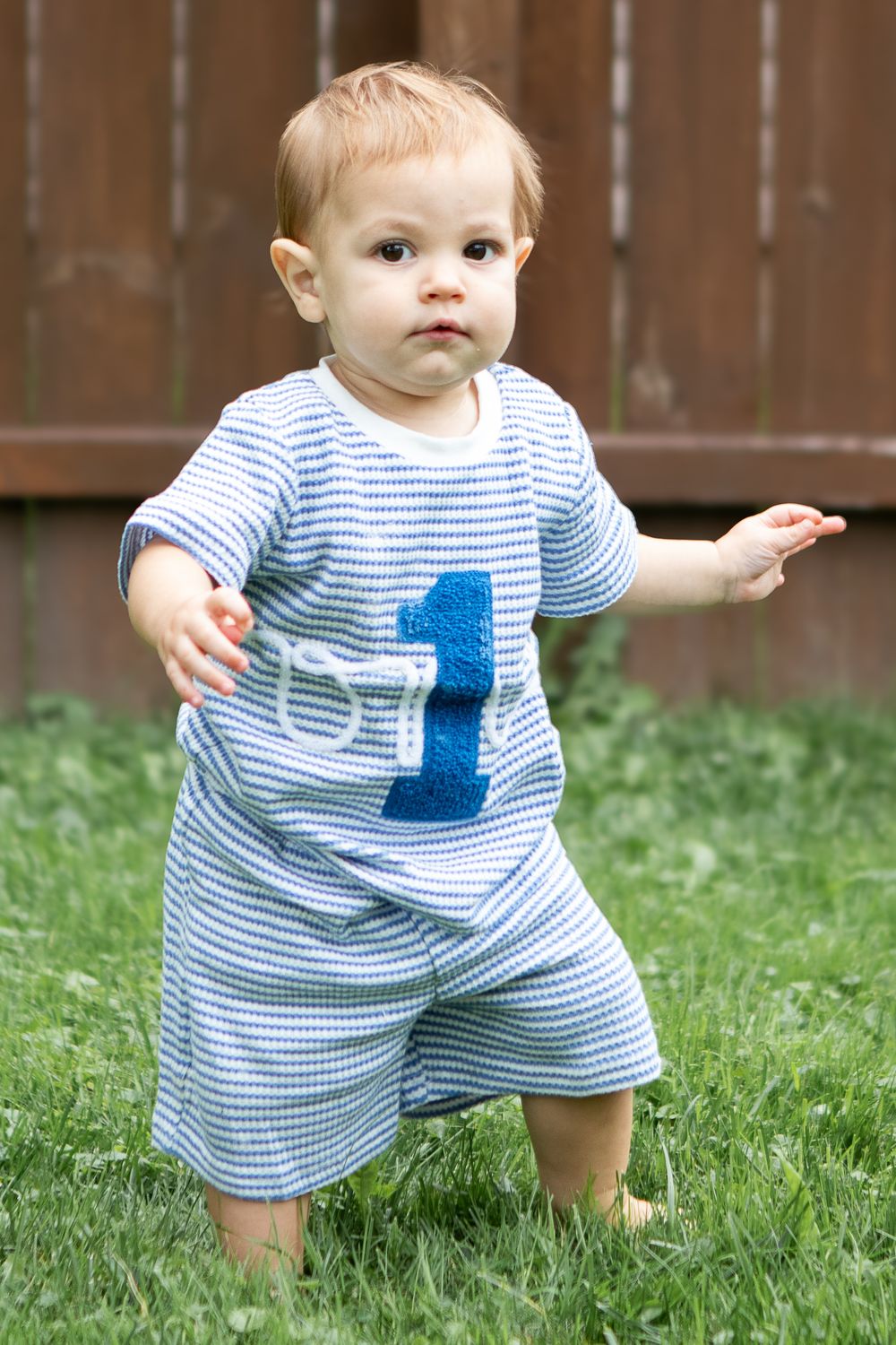 Toddler wearing a blue striped outfit with number 1 patch stands on green grass near a wooden fence.