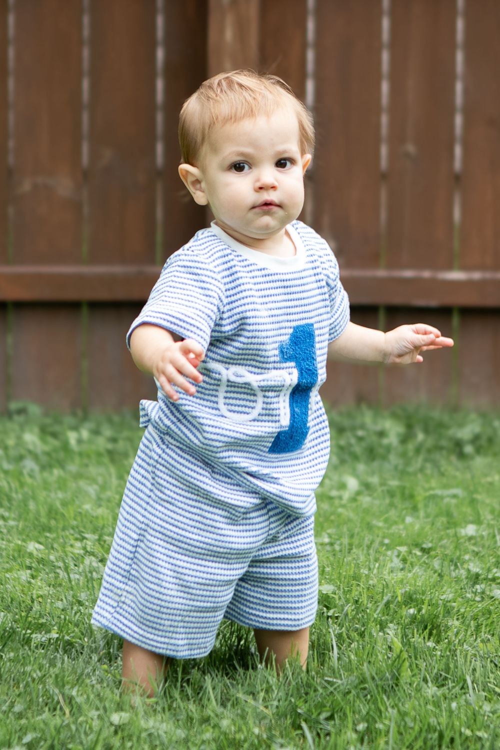 Toddler in blue striped outfit with number 1 standing on green grass in backyard.