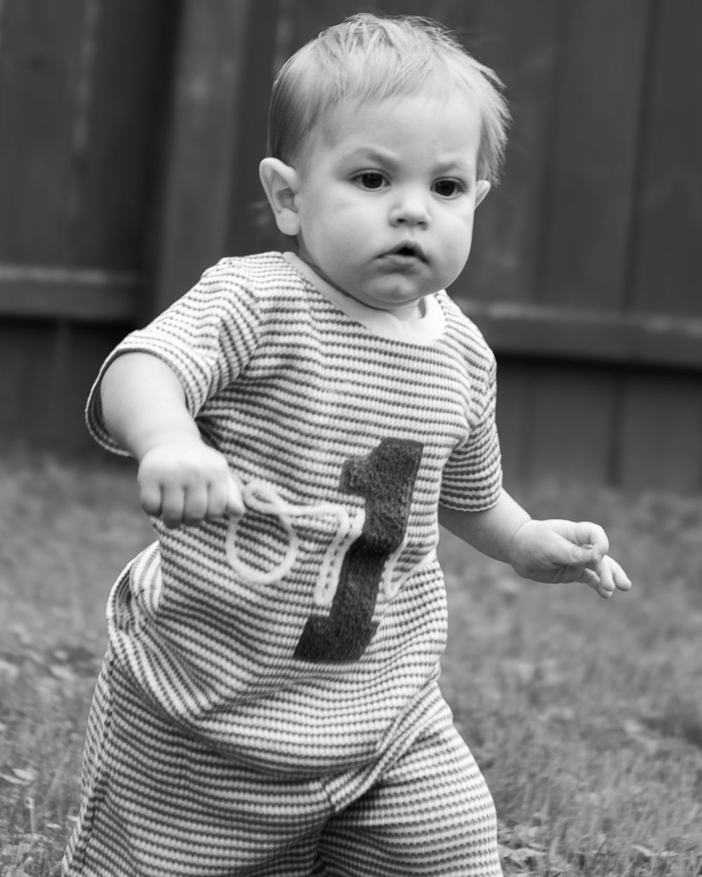 Black and white photo of a toddler wearing a striped romper with the number 1, walking outdoors on grass.