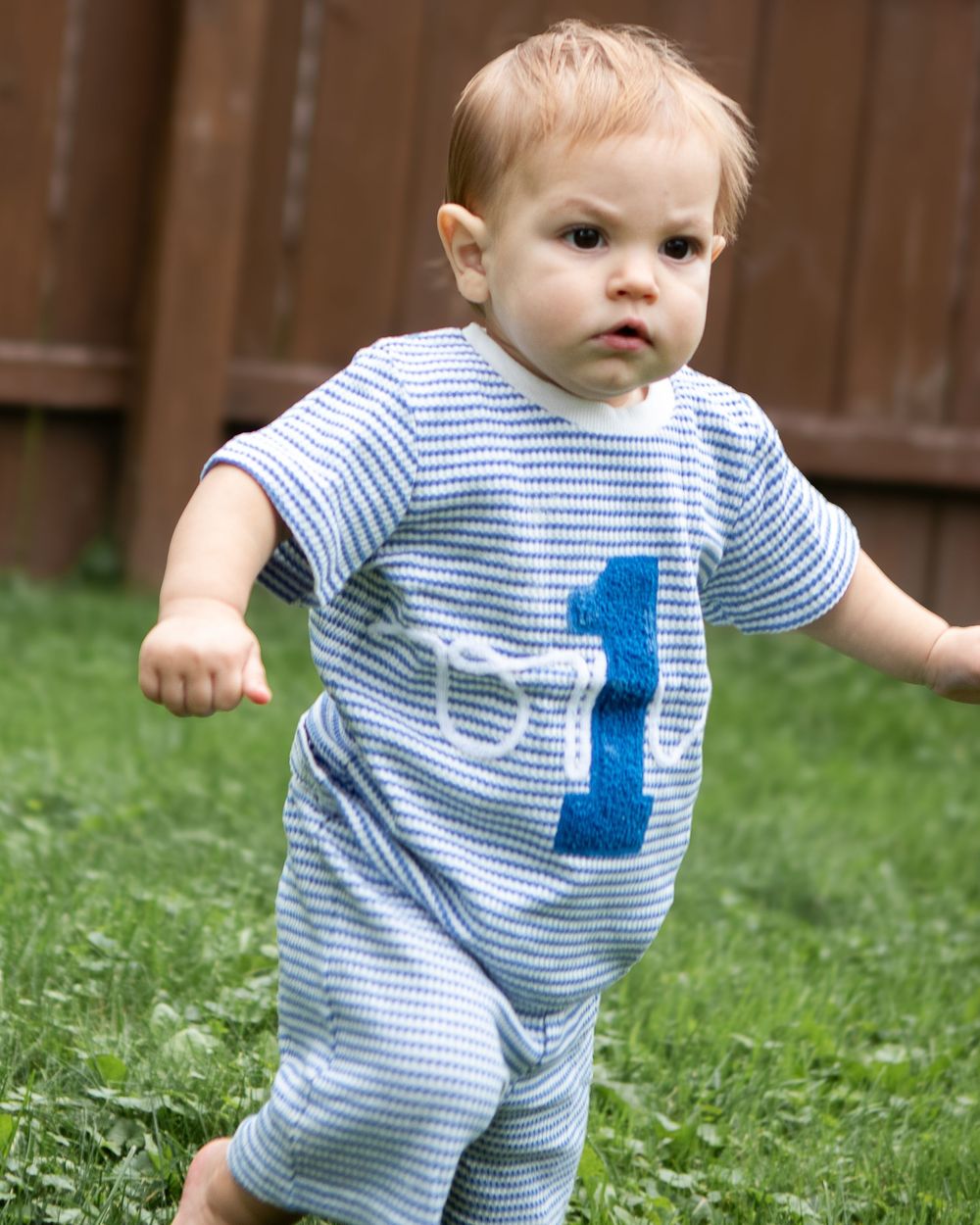 Toddler boy in blue striped number 1 romper walking on green grass for first birthday outdoor photo session.