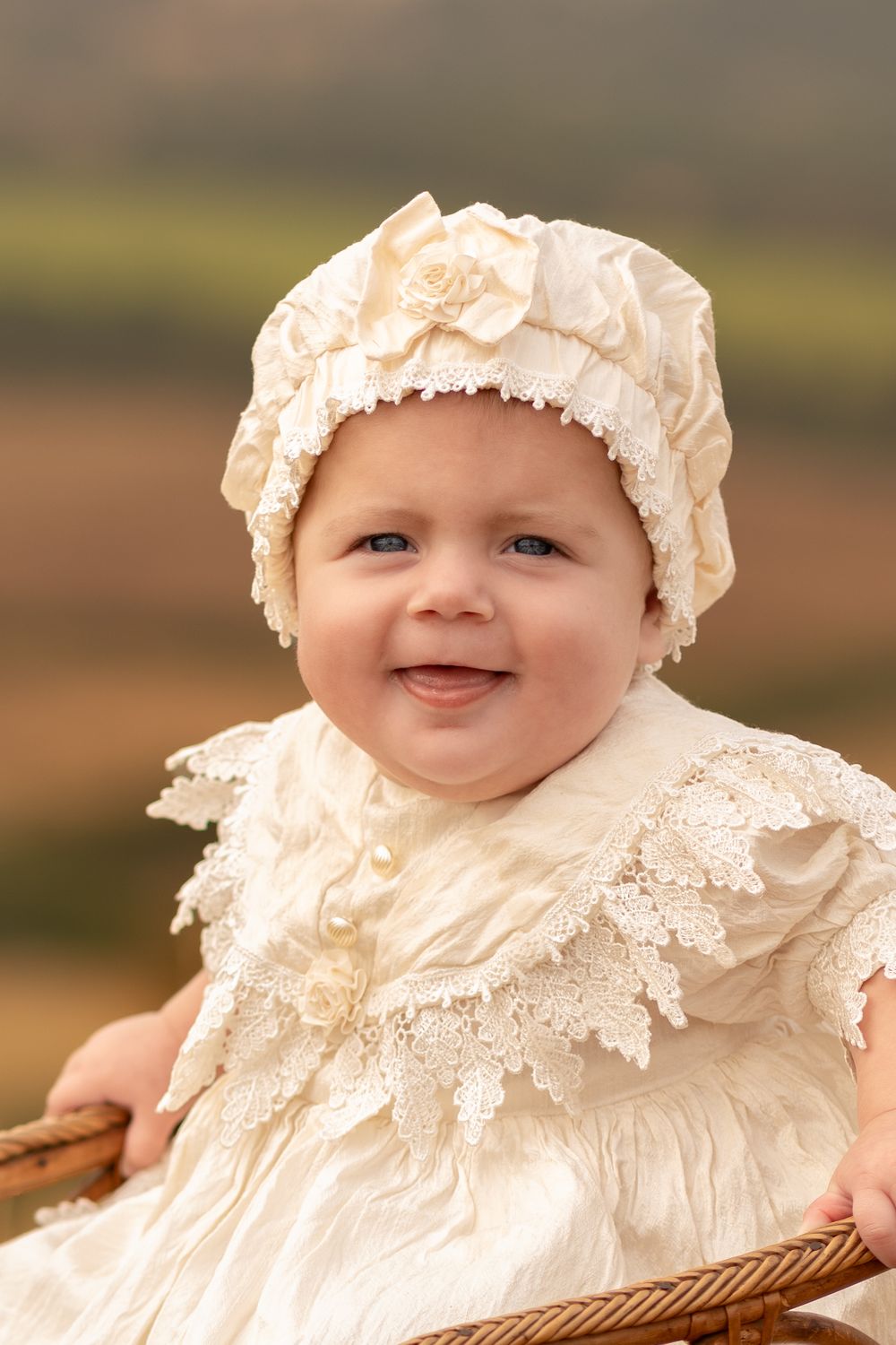 Smiling baby in vintage cream lace dress and bonnet sitting in wicker chair outdoors.