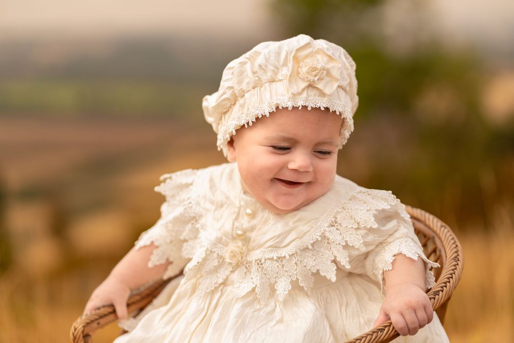 Smiling baby in cream lace dress and bonnet sitting in wicker chair outdoors in golden field.