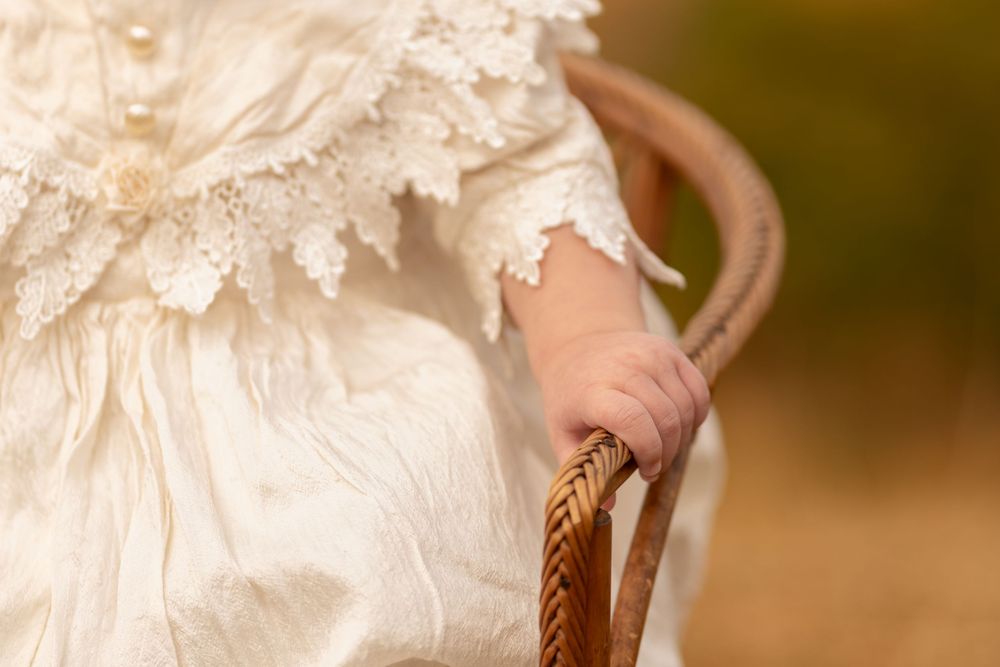 Baby in white lace christening gown holding a wicker basket handle outdoors.