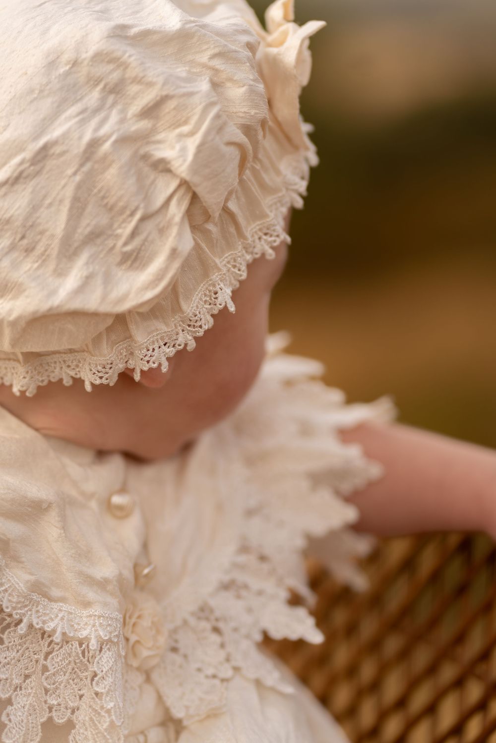 Close-up of a baby wearing an ivory lace christening gown and ruffled bonnet with pearl details.