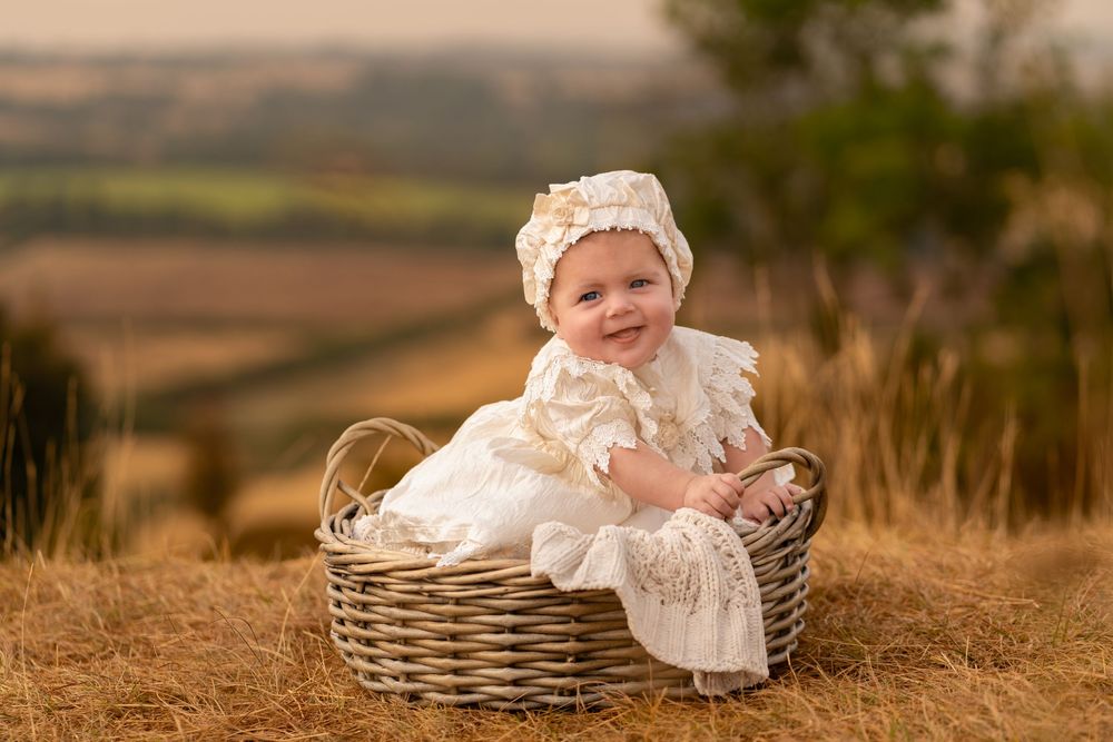 Smiling baby in white lace dress and bonnet sitting in wicker basket outdoors in golden field.