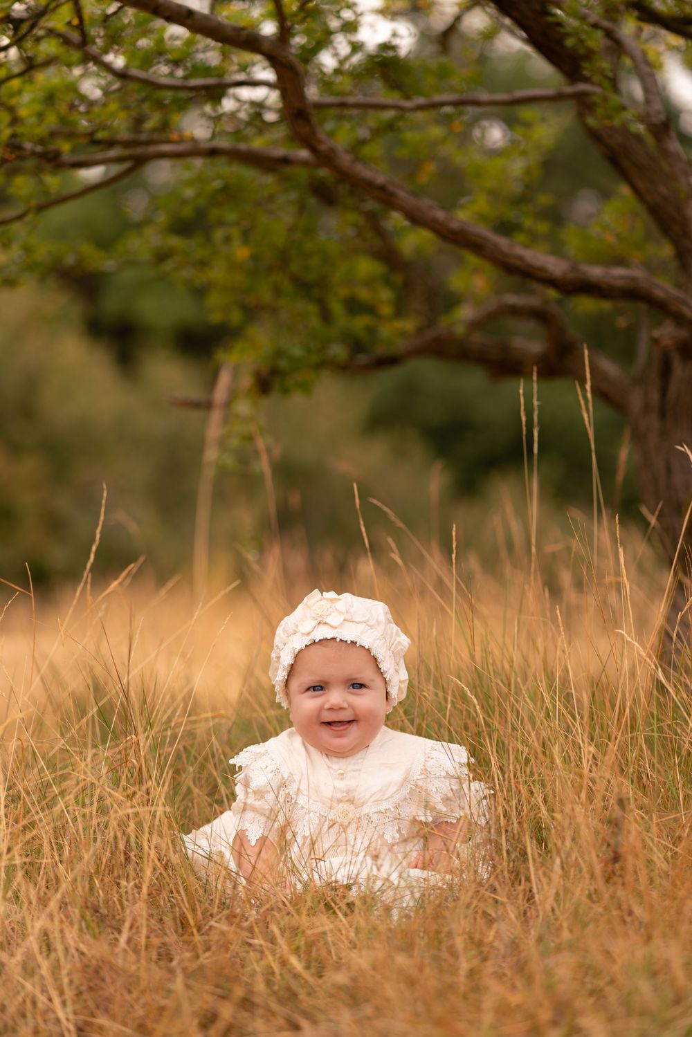 Smiling baby in white dress and bonnet sitting in tall golden grass under a tree outdoors.