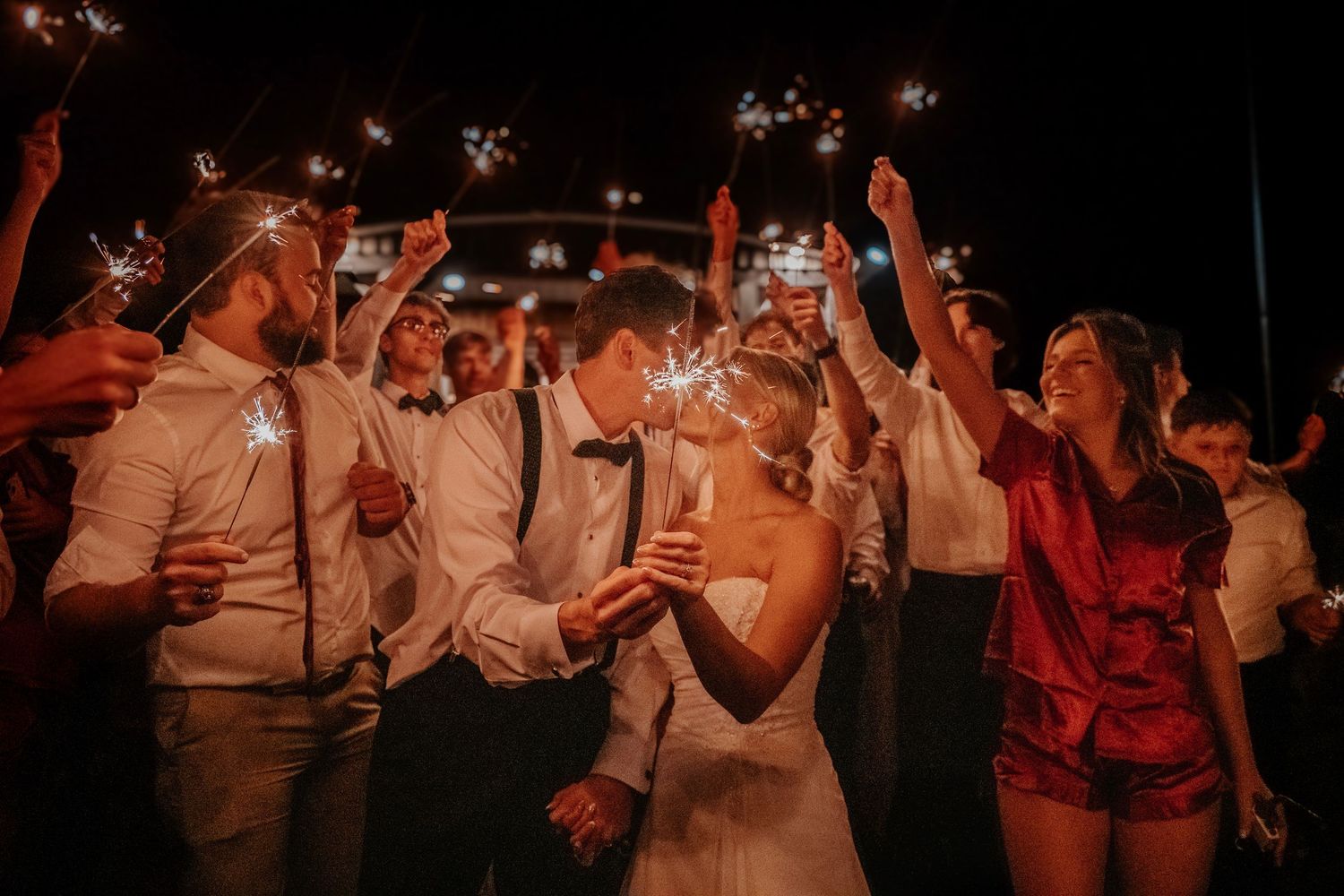 Bride and groom kiss surrounded by wedding guests holding sparklers at a romantic nighttime celebration.