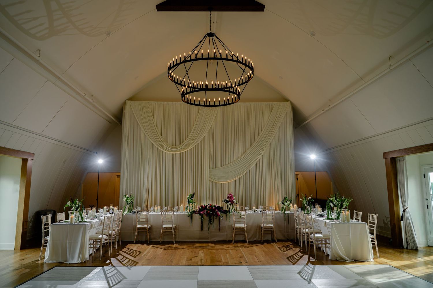 Elegant wedding reception hall with draped white backdrop, round chandelier, and white-clothed tables on hardwood floors.