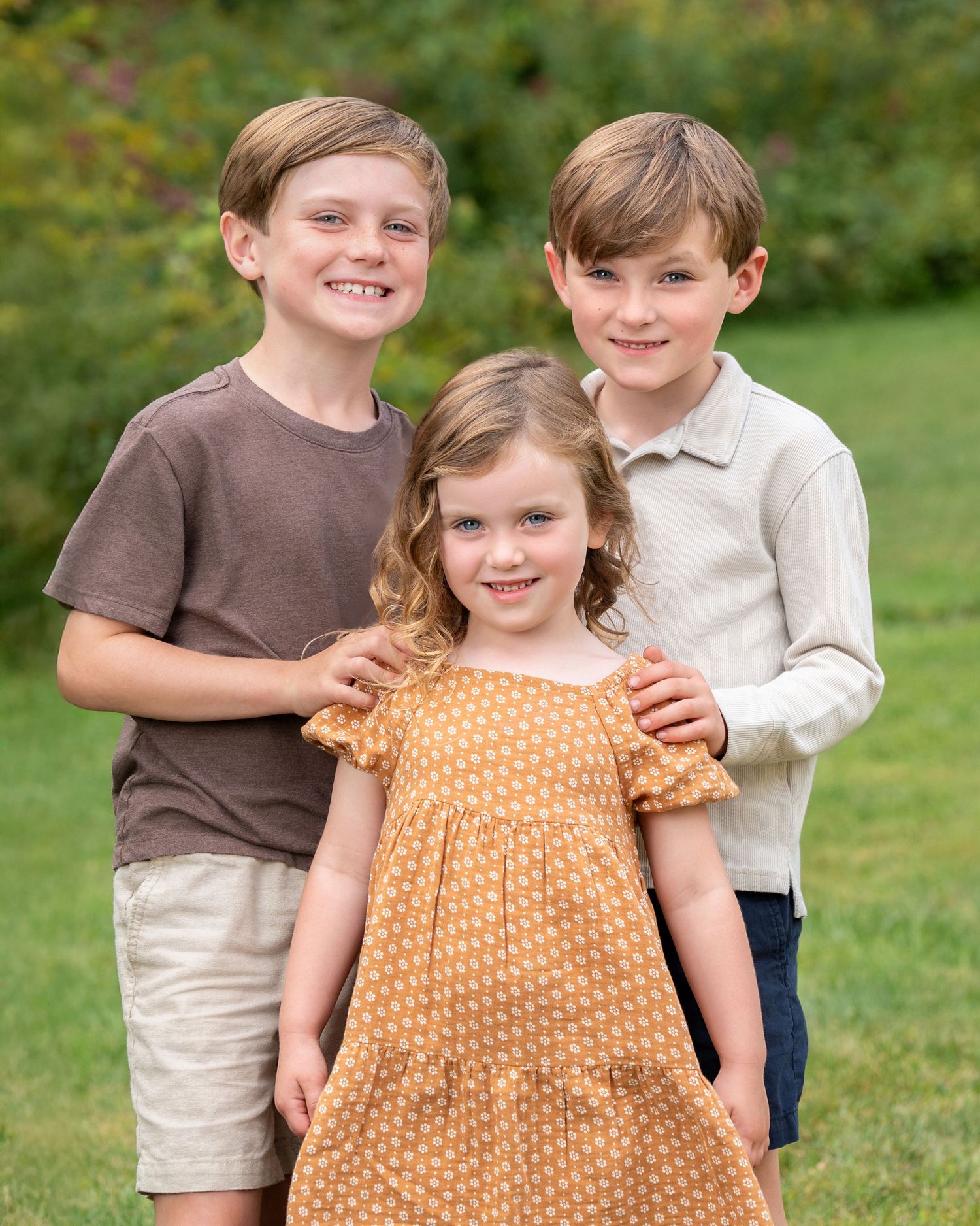 Three smiling children pose outdoors on green grass, two boys flanking a young girl in an orange polka dot dress.