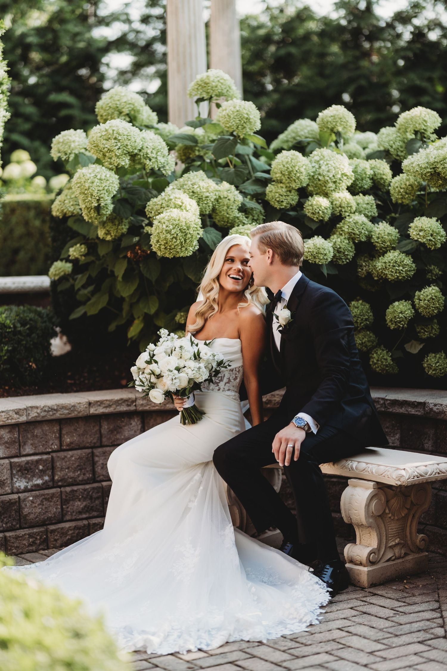 Bride and groom sitting on stone bench laughing, surrounded by lush hydrangea bushes, bride holding white bouquet.