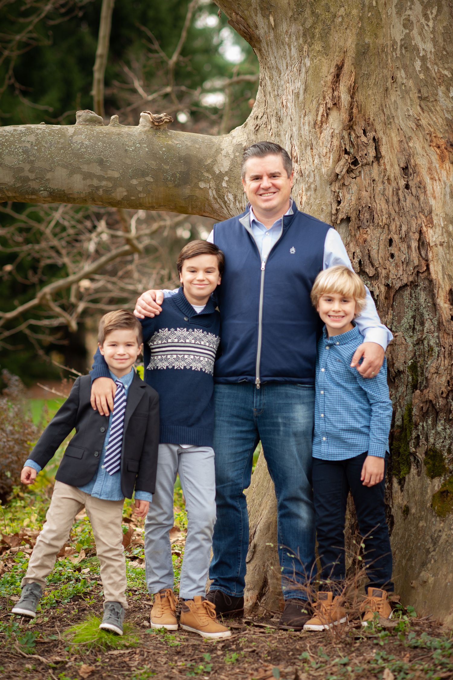 A man and three young boys pose smiling in front of a large tree outdoors in coordinated blue outfits.
