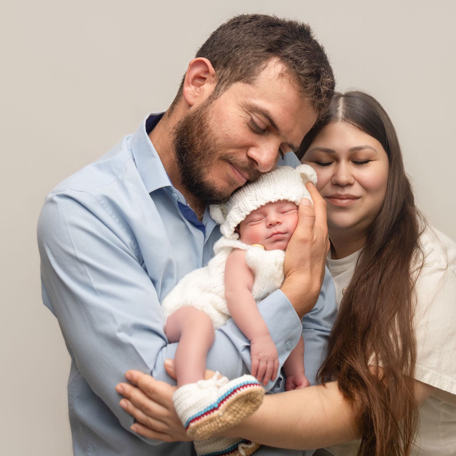A young couple lovingly holds their newborn baby dressed in a white knit outfit against a neutral background.