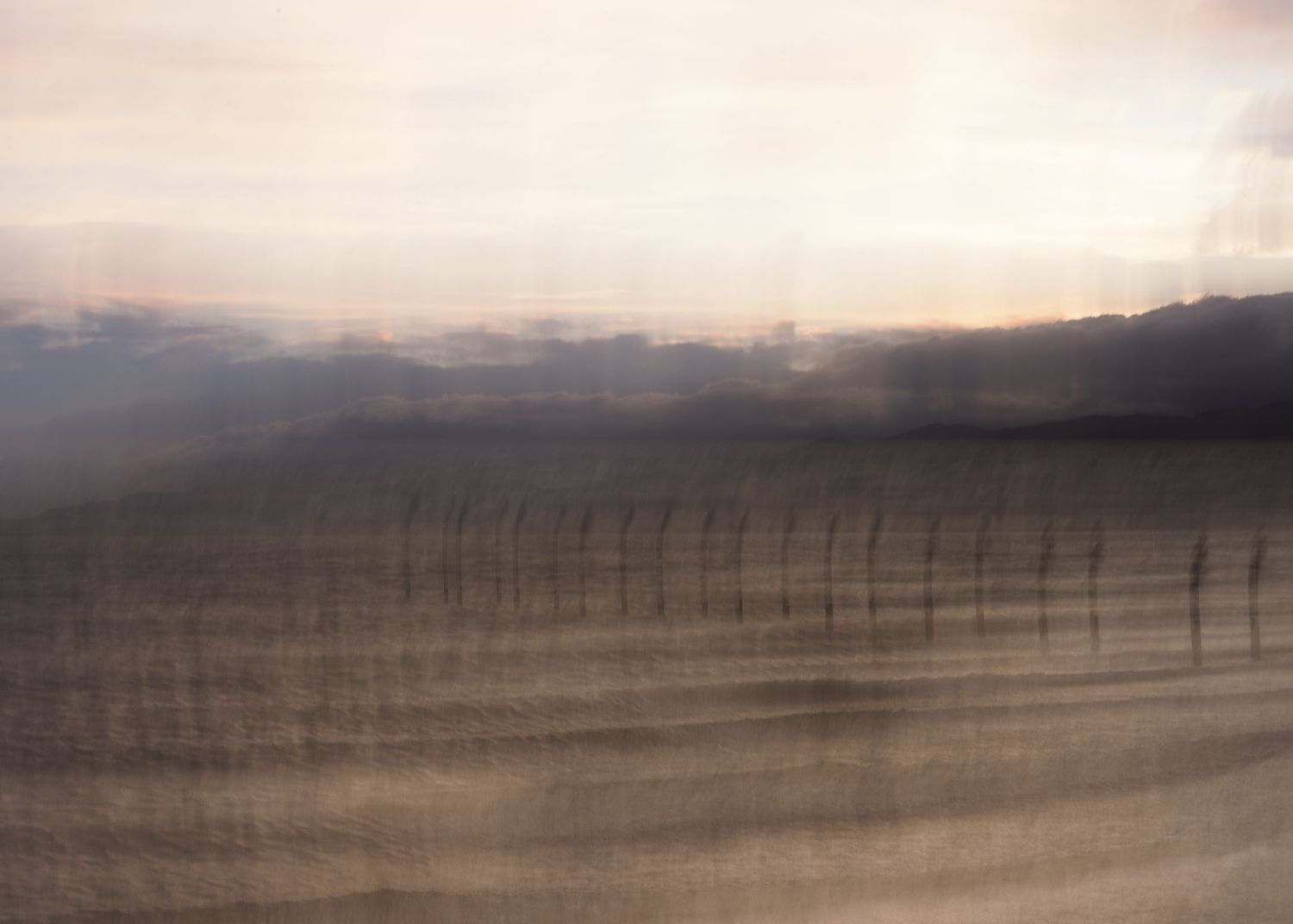 Blurred abstract landscape of misty fields with fence posts at dusk, soft warm tones creating dreamy motion effect.