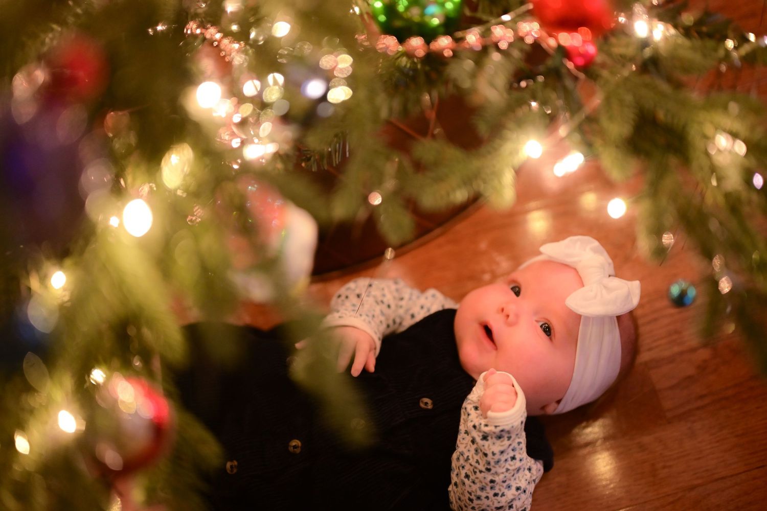 Baby in white bow headband lying under a glowing Christmas tree surrounded by colorful ornaments and lights.