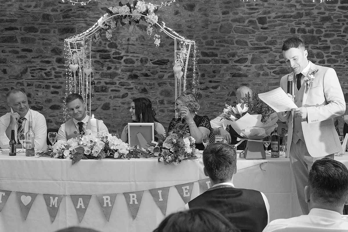 Black and white wedding reception photo showing best man giving speech at head table at New House Farm wedding celebration