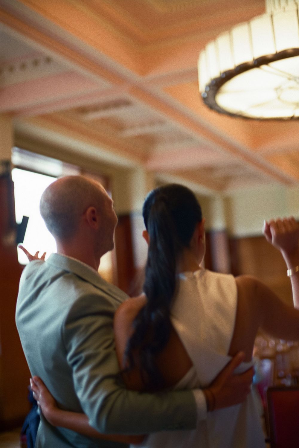 A bald man in a suit embraces a woman with dark hair in a white dress during an elegant indoor event.