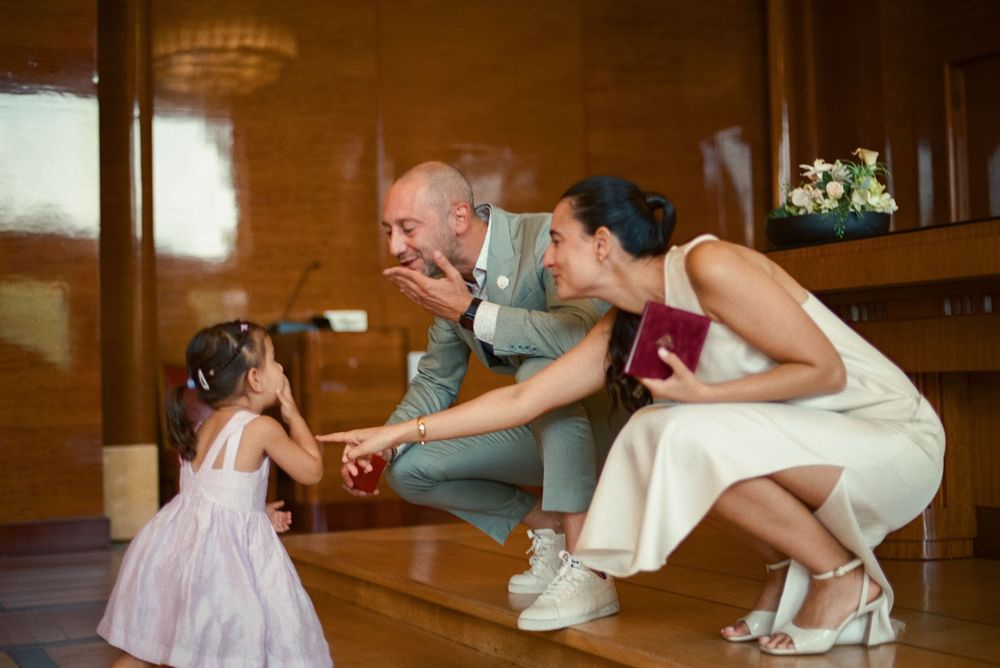 Bride and groom crouching down to interact with a young flower girl during a wedding ceremony indoors.