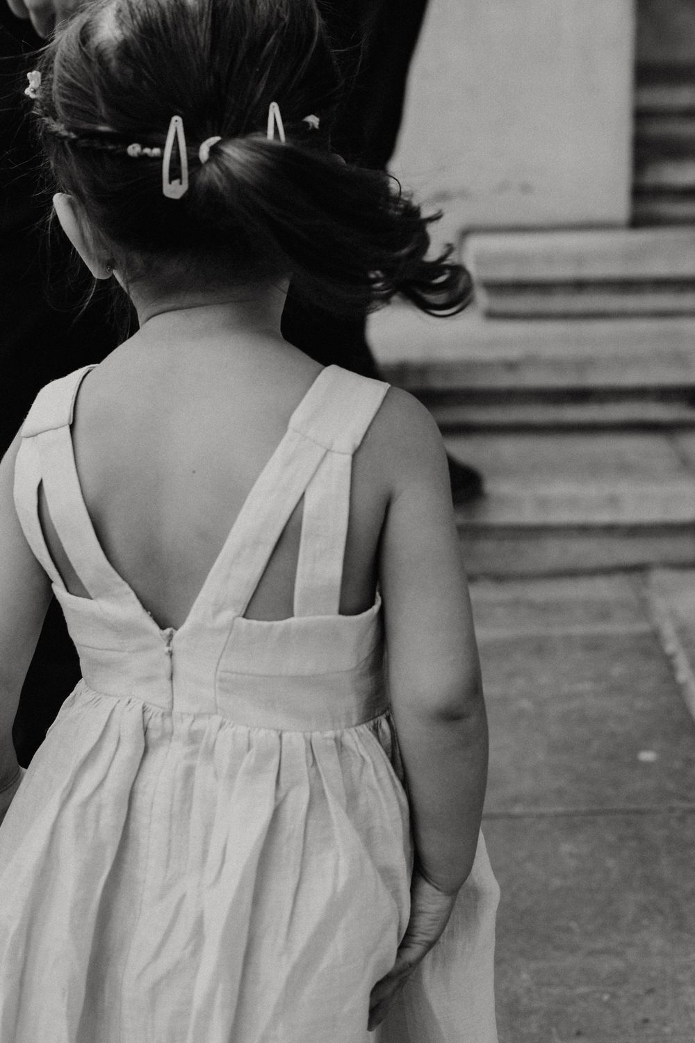 Black and white photo of a flower girl in a white dress with cross-back straps and hair clips at a wedding.