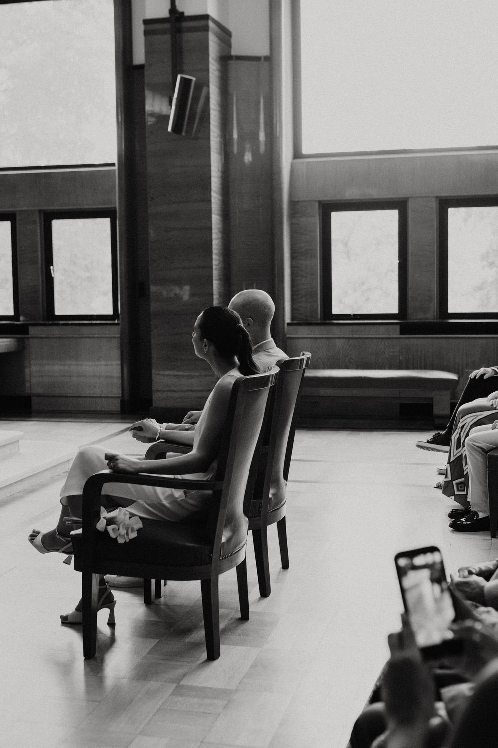 Black and white photo of two people sitting in elegant chairs during a ceremony, facing forward in a bright interior space.