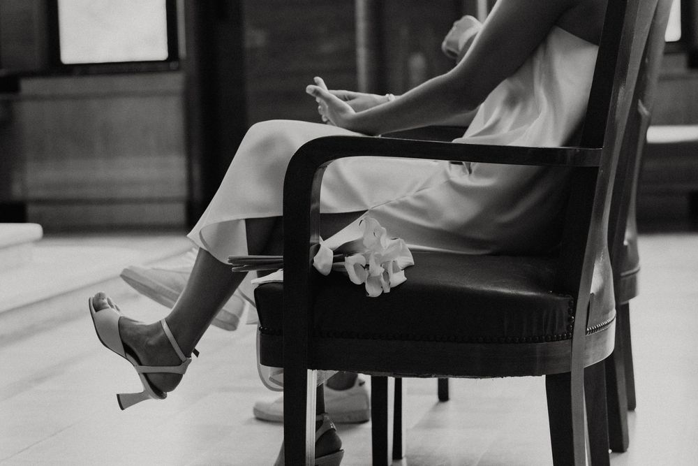 Black and white photo of a woman in a dress and heels sitting elegantly in a chair, holding flowers.