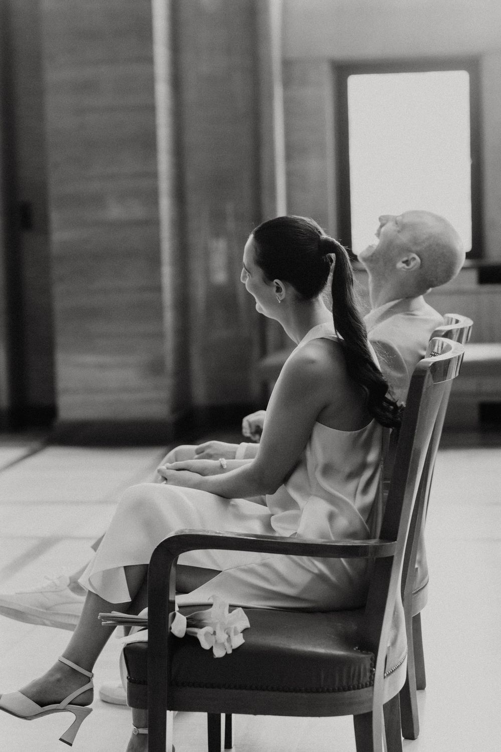 Black and white photo of a bride and groom sitting back-to-back on chairs, sharing a quiet moment before their wedding.