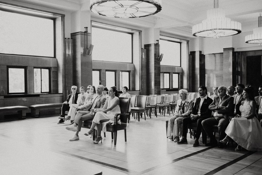 Black and white photo of wedding guests seated in elegant ceremony hall with chandeliers and large windows.