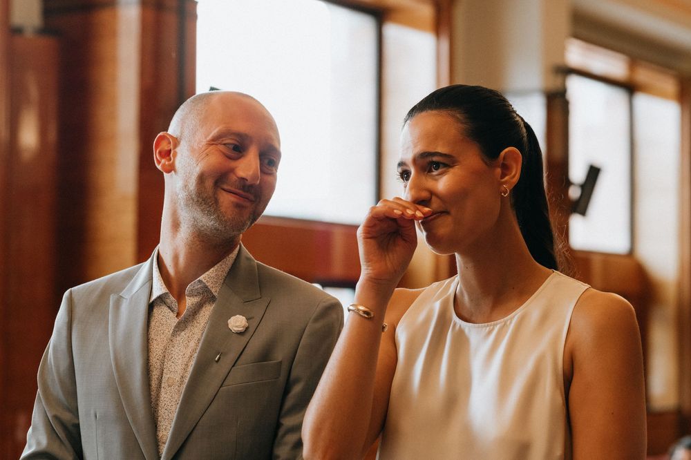 A bald man in a gray suit smiles at a woman in a white dress who covers her mouth, standing indoors.