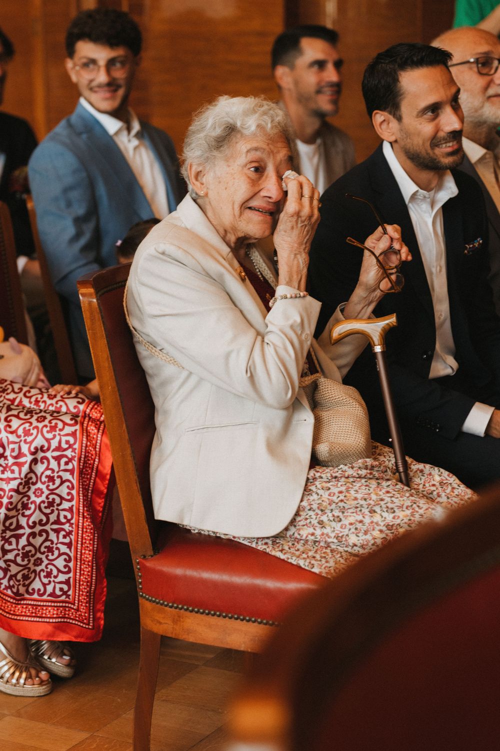 Elderly woman wiping tears at a wedding ceremony, seated among smiling guests in formal attire.