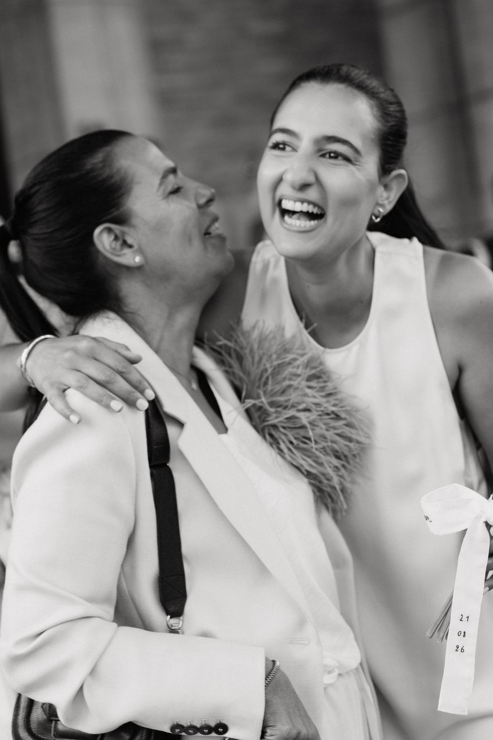 Two women in white outfits laughing and embracing in a joyful black and white photo.