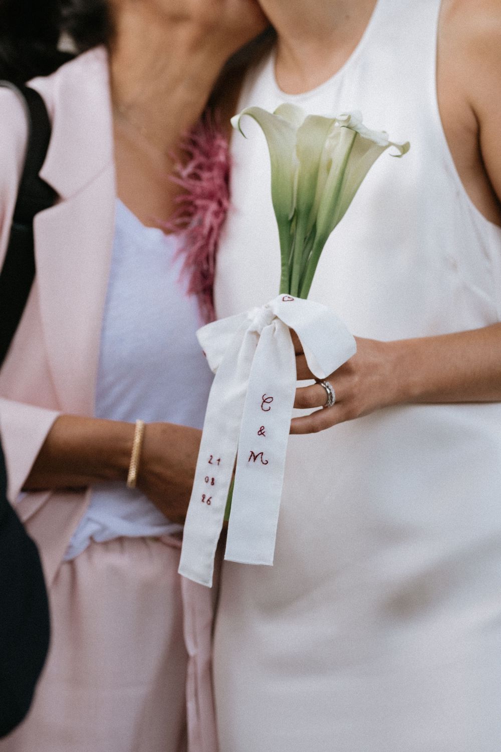 Bride holding white calla lily bouquet with personalized ribbon reading C&M and a wedding date at an outdoor ceremony.