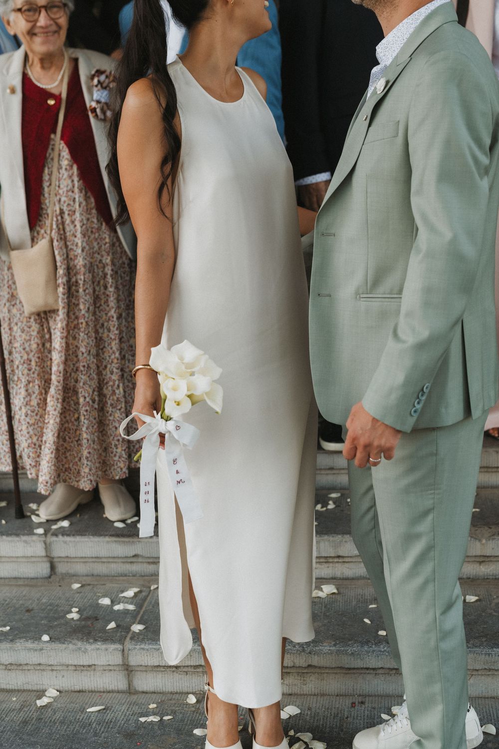 Bride in white midi dress holding calla lily bouquet stands with groom in sage green suit on wedding day.