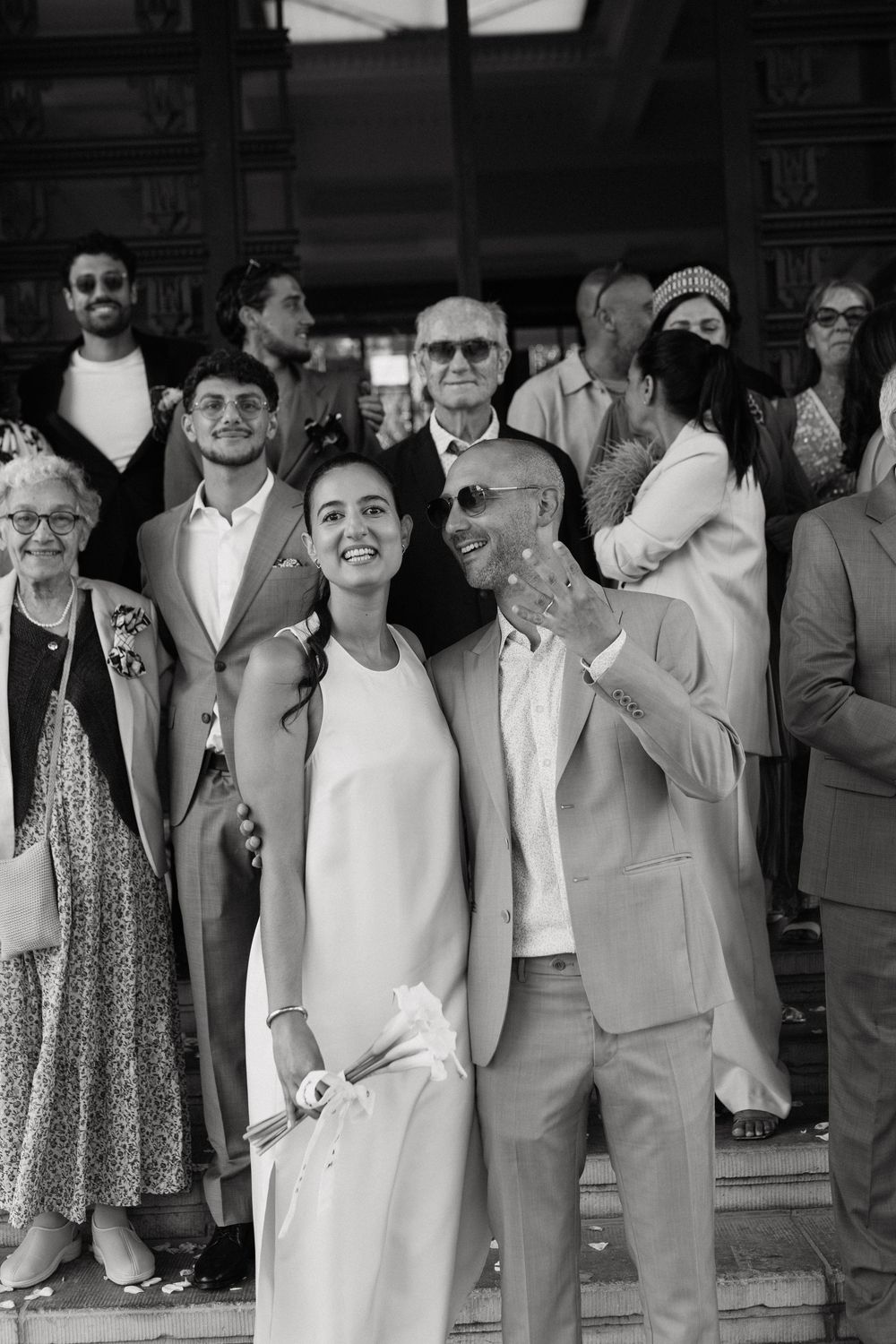 Black and white photo of a smiling bride and groom posing with wedding guests on steps outside a venue.