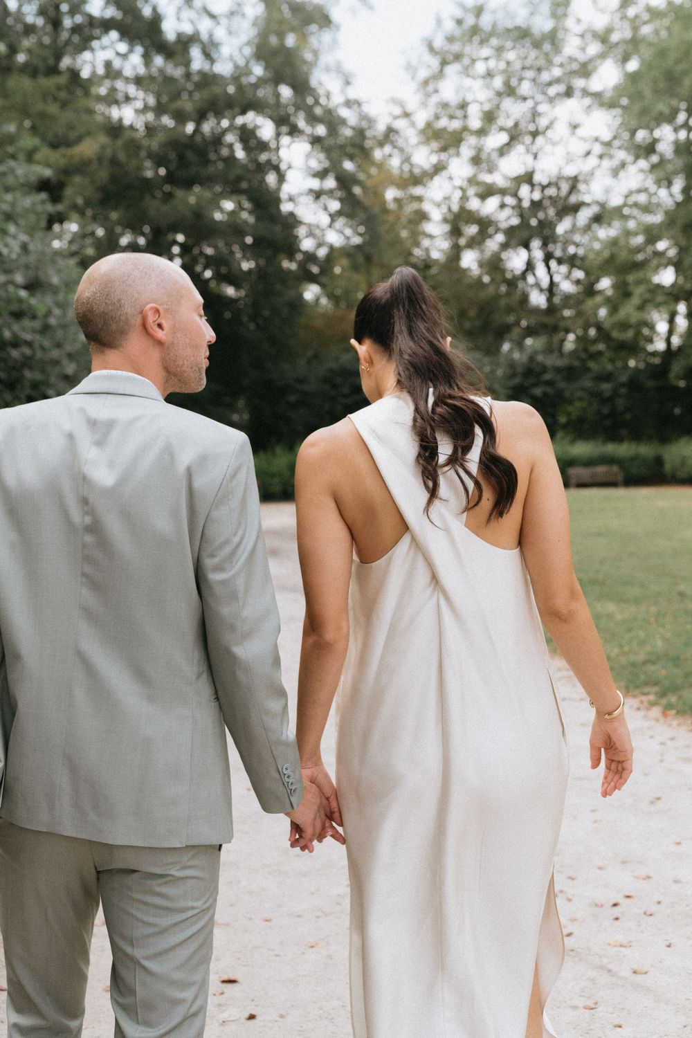 A bride and groom walk hand in hand through a garden, viewed from behind, wearing a white gown and light gray suit.