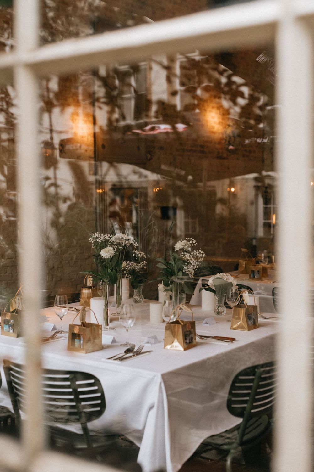 Elegant wedding reception table set with white linens, floral centerpieces, and gold accents, viewed through a window.