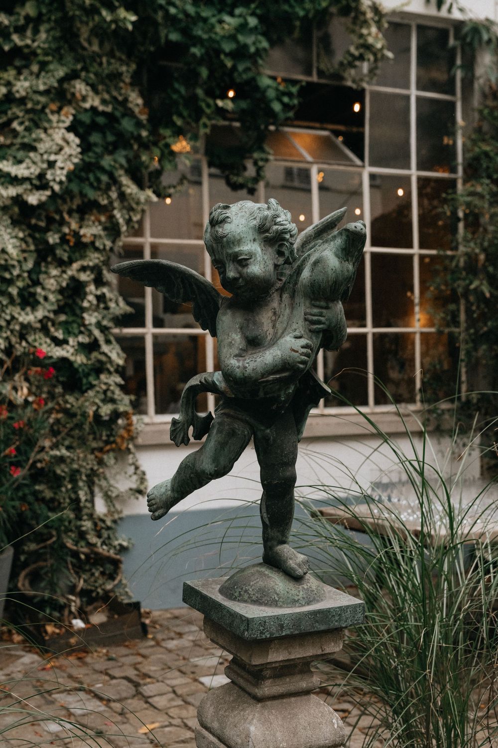 Bronze cherub angel statue on stone pedestal in a lush garden courtyard with ivy-covered walls and large windows.
