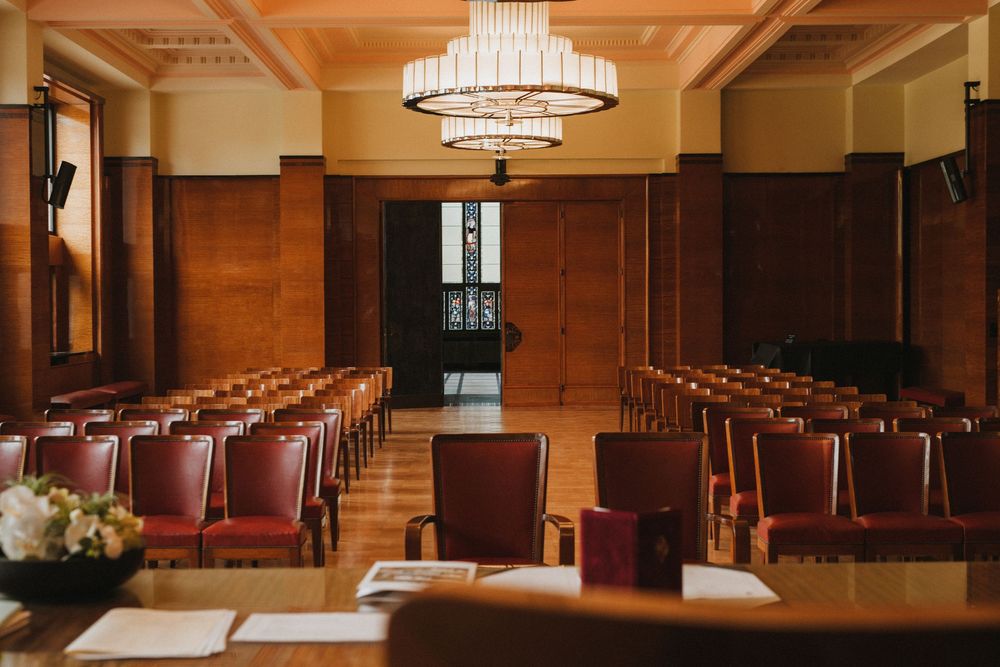 Elegant wood-paneled conference room with red leather chairs, chandelier lighting, and rows of seats in a formal arrangement.